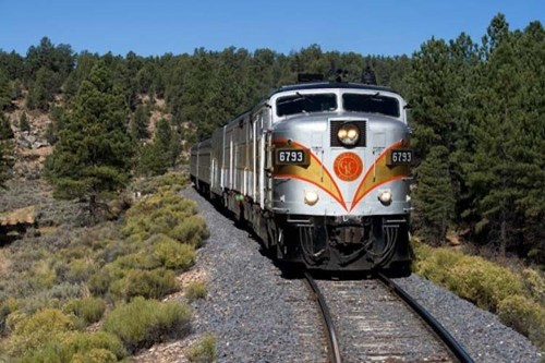 A vintage train travels along a scenic track surrounded by trees and shrubs under a clear blue sky.