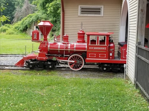 A bright red vintage steam locomotive parked beside a building, surrounded by green grass and trees.