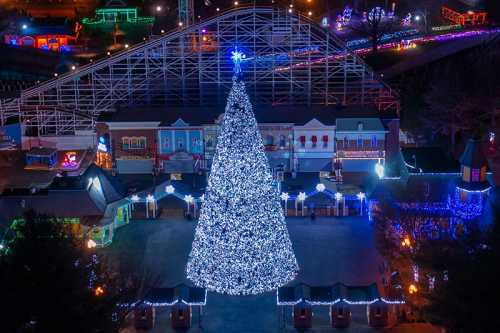 A brightly lit Christmas tree stands in a festive amusement park, surrounded by colorful lights and decorations.