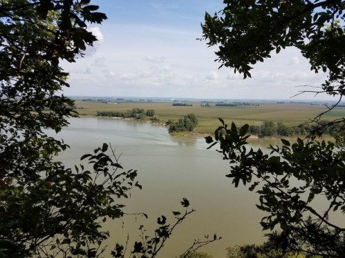 A serene river view framed by green leaves, with fields and distant trees under a partly cloudy sky.
