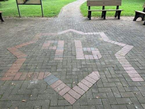 A star-shaped pattern made of bricks on a paved path in a park, with green grass and benches in the background.