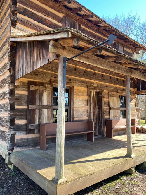 A rustic wooden cabin with a porch, featuring log walls and a simple design, surrounded by trees and blue sky.