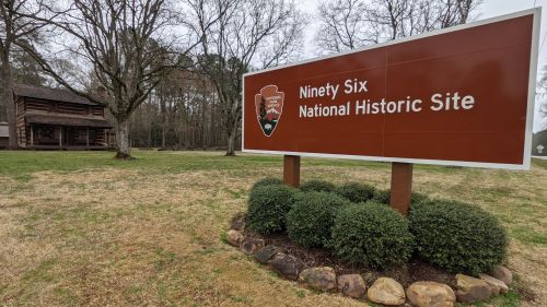 Sign for Ninety Six National Historic Site, with a historic building in the background and trees surrounding the area.