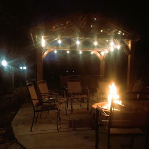 A cozy outdoor gazebo at night, illuminated by string lights, with chairs around a fire pit.