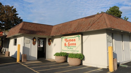 Exterior of Tumea & Sons Italian Restaurant and Lounge, featuring a sign and planters, under a blue sky.
