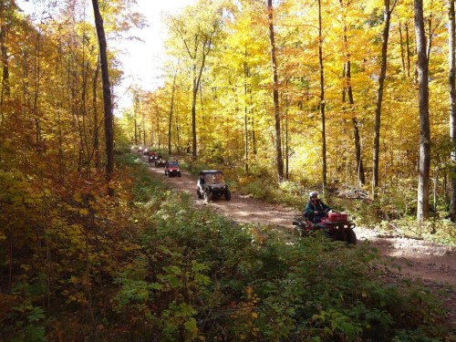 A line of off-road vehicles travels down a dirt path through a vibrant autumn forest.