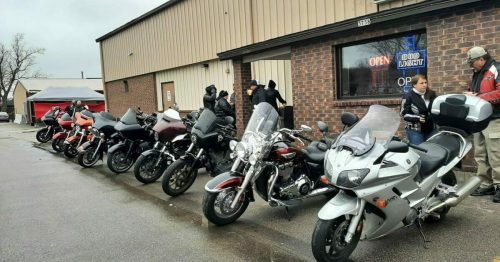 A row of various motorcycles parked outside a sports center, with people gathered nearby.