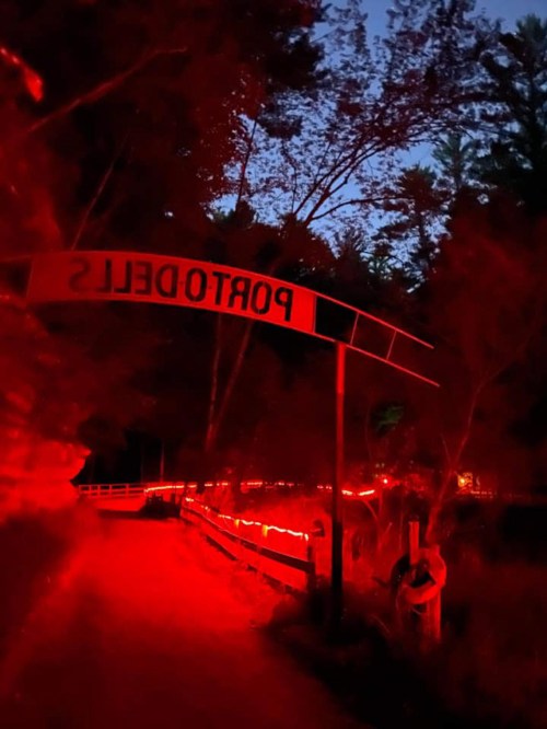 A dimly lit path with red lights and a sign reading "PORTODLETIS" surrounded by trees at dusk.