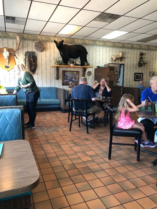 A cozy restaurant interior with diners at tables, a girl sitting with adults, and a bear statue on a shelf.