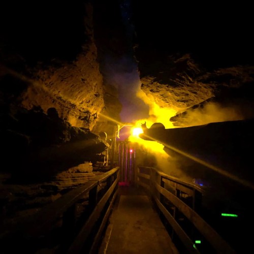 A dimly lit cave with a wooden pathway, illuminated by warm yellow and blue lights, creating a mystical atmosphere.