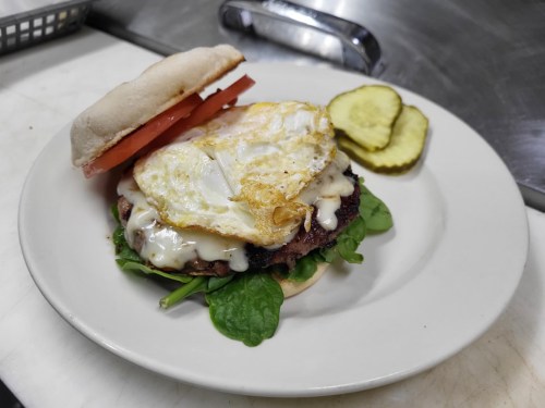 A burger with spinach, tomato, and a fried egg, topped with creamy sauce, served with pickles on the side.