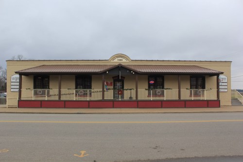 A one-story building with a metal roof, featuring a porch and large windows, located along a road.