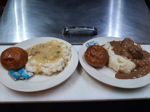 Two plates of mashed potatoes topped with gravy and vegetables, accompanied by small bread rolls and butter packets.
