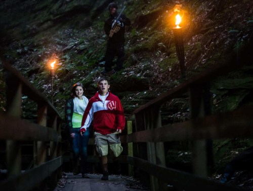 A couple walks hand in hand on a bridge, while a figure with a weapon stands in the background, illuminated by torches.
