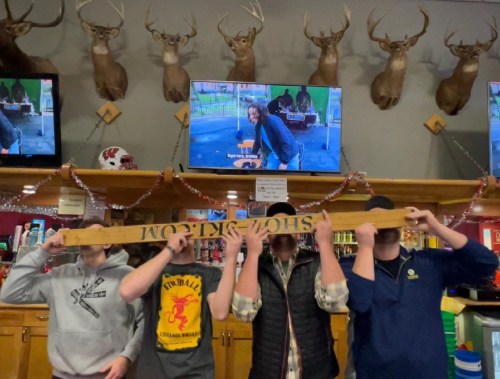 Four people hold a long wooden sign in front of their faces, with deer heads mounted on the wall and screens in the background.