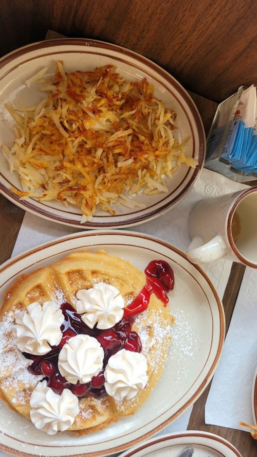 A plate of waffles topped with whipped cream and cherry sauce, alongside a serving of crispy hash browns.
