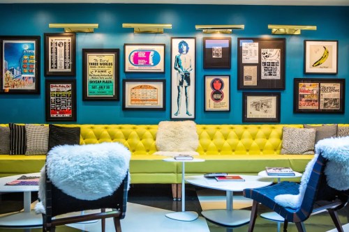 A vibrant lounge area with a yellow tufted sofa, framed art on blue walls, and modern chairs around white tables.