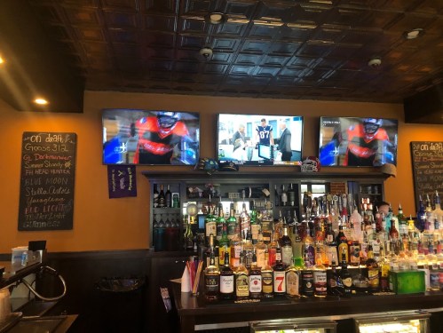 A bar with a wooden counter, shelves of liquor, and three TVs showing sports and news. A chalkboard lists drink specials.
