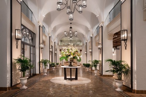 Elegant hallway with arches, decorative lighting, potted plants, and a central floral arrangement on a round table.