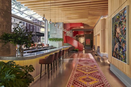 Modern hotel lobby featuring a sleek bar, colorful artwork, and a patterned rug under wooden beams and large windows.