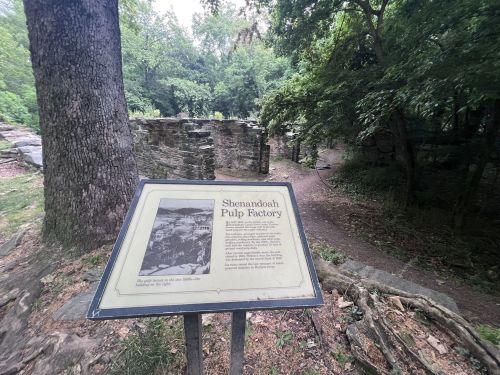 Sign for the Shenandoah Pulp Factory, surrounded by trees and ruins, with a path leading into the forest.
