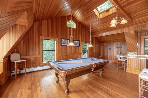 A wooden-paneled game room featuring a pool table, bar area, and large windows for natural light.