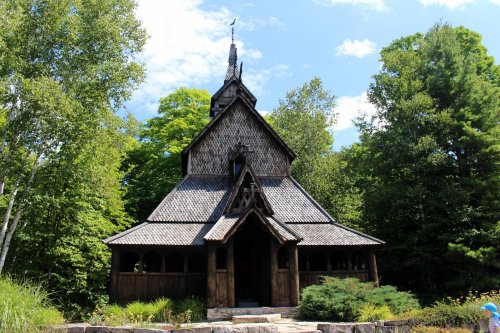 A wooden church with a steep roof, surrounded by lush green trees and a blue sky.