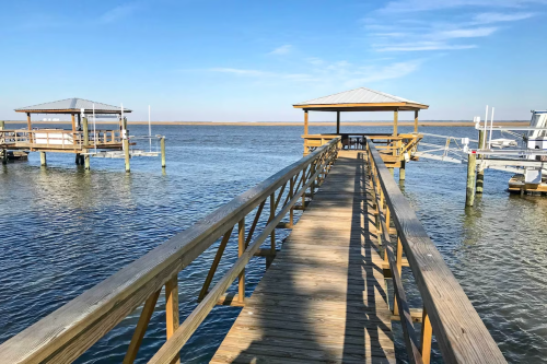 A wooden dock extends over calm water, leading to two covered platforms under a clear blue sky.