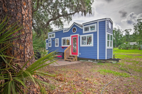 A blue tiny house with a red door, surrounded by greenery and trees, set in a spacious outdoor area.