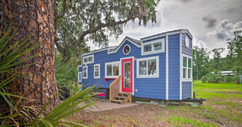 A small blue tiny house with red accents, surrounded by greenery and trees, featuring a porch and large windows.