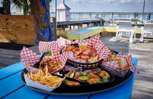 A colorful seafood platter with shrimp, fries, and sides, set on a table by a waterfront pier.