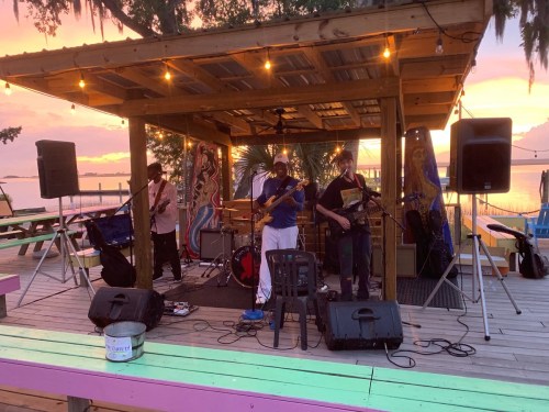 A band performs on a colorful outdoor stage at sunset, with lights and palm trees in the background.