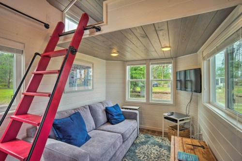 Cozy interior of a tiny house featuring a gray sofa, blue pillows, a wooden table, and a red ladder leading to a loft.