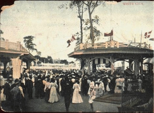 A vintage postcard depicting a crowded amusement park scene with people in period clothing and flags overhead.