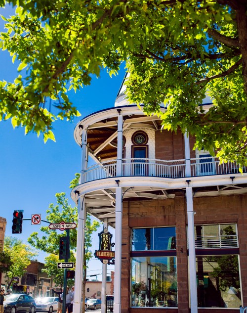 A sunny street view of a historic building with a curved balcony, surrounded by green trees and clear blue skies.