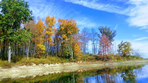 A serene lakeside scene with colorful autumn trees reflecting in the water under a blue sky with scattered clouds.