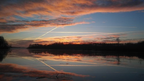 A serene sunset over a calm river, with colorful clouds and reflections, intersected by contrails in the sky.