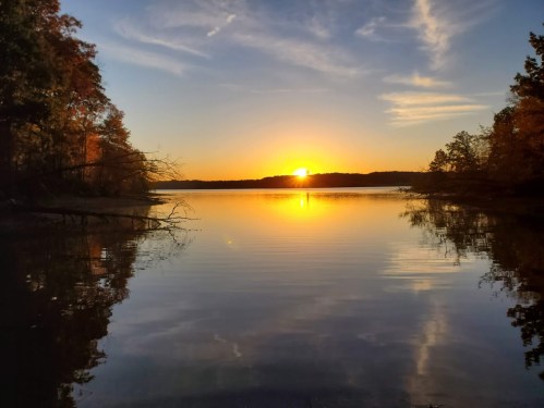 A serene lake at sunset, with vibrant colors reflecting on the water and trees lining the shore.