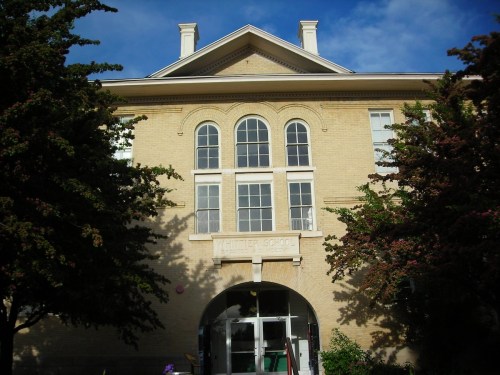 Historic school building with arched entrance, large windows, and trees in front under a blue sky.