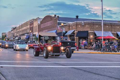 A vintage red car drives through a bustling street with shops and people enjoying the evening atmosphere.