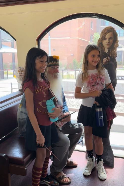 Two girls pose with a man in a wizard costume on a train, while a cardboard cutout of a female character stands nearby.