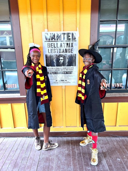 Two girls in wizard costumes pose playfully in front of a "Wanted" poster for Bellatrix Lestrange.
