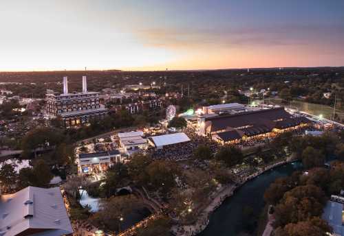 Aerial view of a lively outdoor event near a river at sunset, featuring a ferris wheel and large gathering spaces.