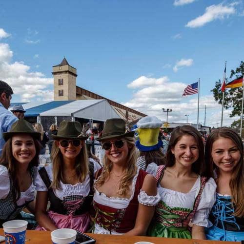 A group of five women in traditional attire smiling at a festival, with a building and flags in the background.