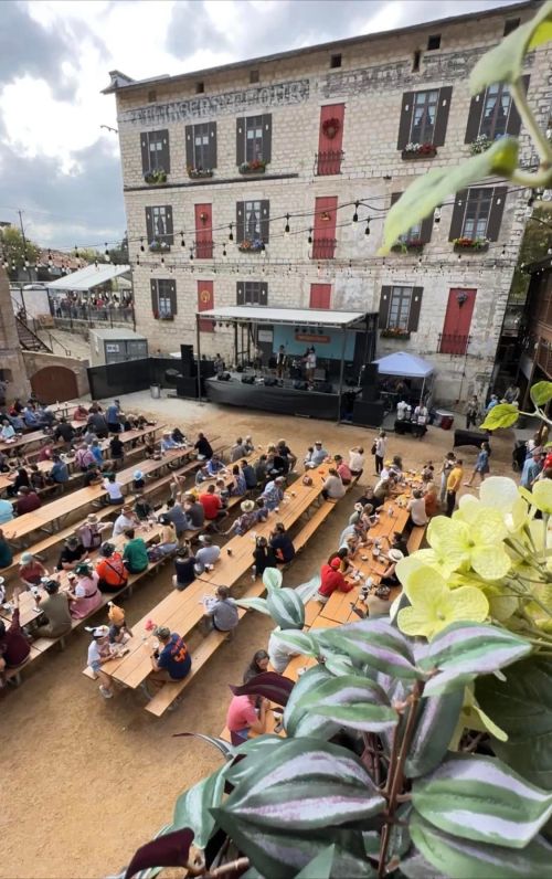 A lively outdoor event with a stage, audience seated at long tables, and a charming stone building in the background.