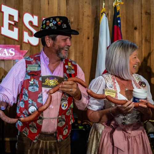 Two people in traditional attire smile and hold sausages at a festive event, with flags and wooden decor in the background.