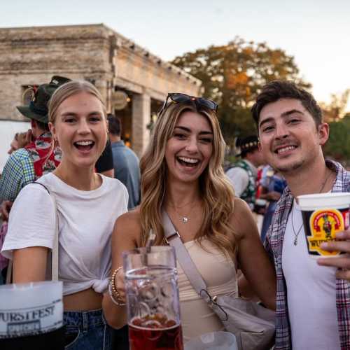 Three smiling friends pose together at an outdoor festival, holding drinks with a lively crowd in the background.