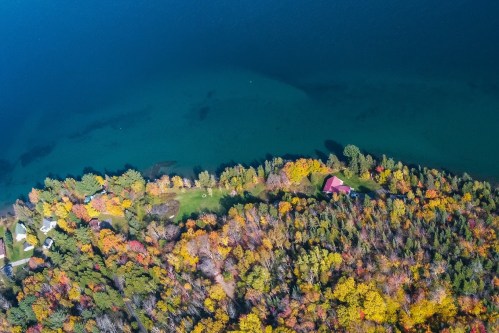 Aerial view of a colorful forest in autumn, with a red-roofed house near a blue lake.