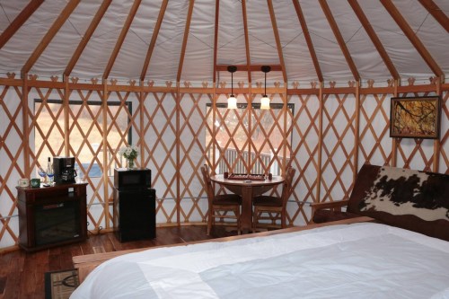 Interior of a cozy yurt featuring a bed, dining table, and kitchenette with wooden accents and large windows.
