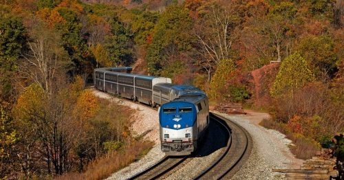 A train curves through a colorful autumn landscape with trees in shades of orange, yellow, and green.